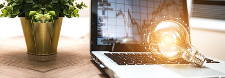 Spotmarkt und Terminmarkt: So funktioniert die Strombörse Ein Notebook, auf dem Börsenkurse zu sehen sind und auf dessen Tastatur eine Glühbirne liegt, auf einem Schreibtisch, daneben eine grüne Topfpflanze