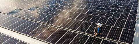 Pflichten für PV-Anlagen-Betreiber Ein PV-Modul-Feld auf einem Hallendach, darauf ein Mann mit Helm und Meßgerät