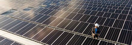 Pflichten für PV-Anlagen-Betreiber Ein PV-Modul-Feld auf einem Hallendach, darauf ein Mann mit Helm und Meßgerät