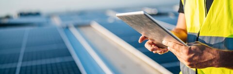 Photovoltaik Checkliste Ein Mann in gelber Warnweste hält ein Tablet in den Händen, im Hintergrund eine PV-Anlage