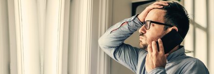 Aktuelle Trends der Cyberkriminalität Ein Mann steht im Büro, schaut durch das Fenster nach draußen, telefoniert mit einem Smartphone und langt sich mit seiner rechten Hand an den Kopf.