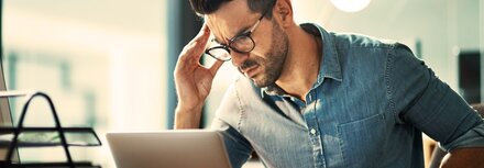 BSI-Bericht: Die Zahl der Cyberangriffe hat sich erhöht. Ein Mann sitzt am Schreibtisch vor einem Laptop, reibst sich mit seiner rechten Hand die rechte Schläfe, grübelt über einen erfolgreichen CEO-Fraud-Angriff auf sein Unternehmen.