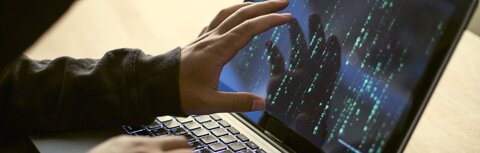 DDoS-Attacken Ein Hacker sitzt an einem Laptop und zeigt mit seiner Hand auf den Bildschirm. Auf dem Bildschirm sind Zahlen und Buchstaben zu sehen.