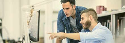 Managed Security von EWE Zwei junge Männer am Schreibtisch in einem Büro. Einer der Männer sitzt auf seinem Stuhl und zeigt mit seinem Arm auf seinen Monitor. Der andere Mann steht neben ihm und schaut auf den Monitor.