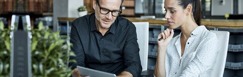 Bild: Morsa Images / DigitalVision / Getty Images Eine Frau und ein Mann sitzen an einem Schreibtisch in einem Büro und schauen sich etwas auf einem Tablet-PC an.