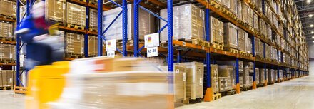 Flächenmanagement mit Licht für Industriehallen Ein Gabelstapler, der nur verschwommen erkennbar ist und gerade um die Ecke in einer gut beleuchteten Lagerhalle fährt
