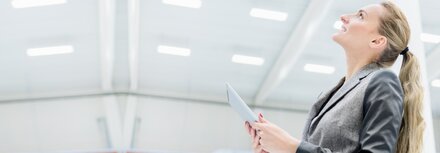 Licht-Monitoring mit EWE Eine blonde Frau im grauen Anzug blickt nach oben und überprüft begeistert die Lichtsteuerung per Tablet.