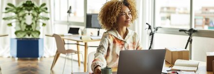 Die Zukunft des Wohnens: Quartiere für die Generation Z Eine Frau sitzt an einem Schreibtisch in einem Bürogebäude und guckt in die Ferne, im Hintergrund lehnt ein Fahrrad an der Fensterfront.
