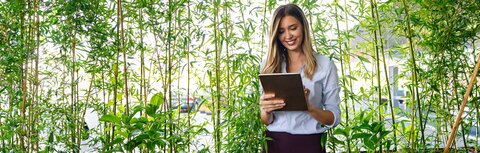 Freundlich zum Klima Eine Frau mit Tablet steht vor einer Reihe Pflanzen, im Hintergrund sind ein Firmengebäude und PKW zu sehen.