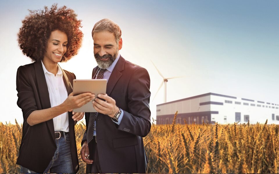 Eine Frau und ein Mann stehen in einem Getreidefeld vor einer Fabrik mit Windkraft und gucken begeistert auf ein Tablet.