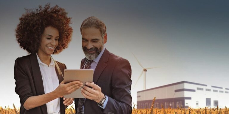 Eine Frau und ein Mann stehen in einem Getreidefeld vor einer Fabrik mit Windkraft und gucken begeistert auf ein Tablet.