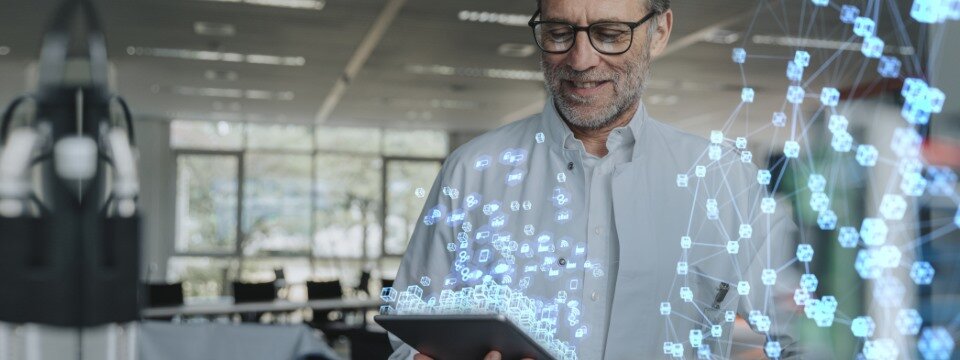 Ein Mann guckt in einem Büro auf ein Tablet, auf dem eine stilisierte hellblaue Vernetzung mit der Umgebung angedeutet wird.
