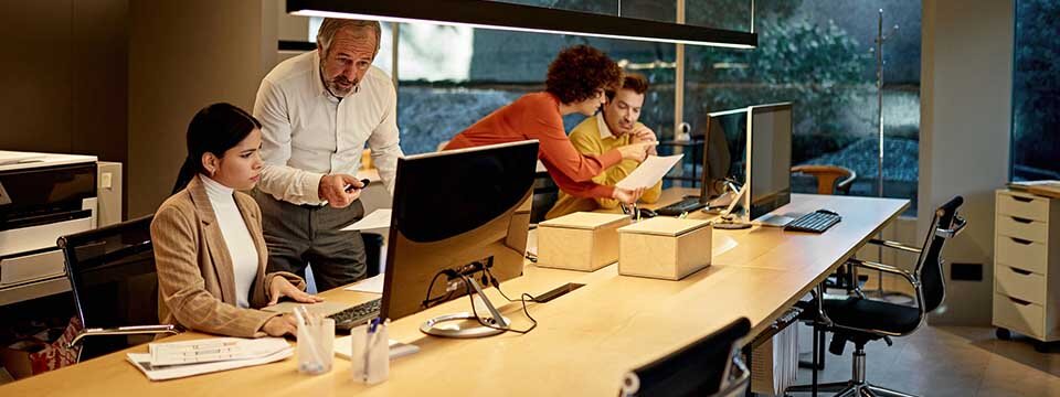 Zwei Mitarbeitende in einem Büro vor Rechner werden von zwei älteren Mitarbeitenden unterstützt, die hinter ihnen stehen. | © Bild: xavierarnau / E+ / Getty Images