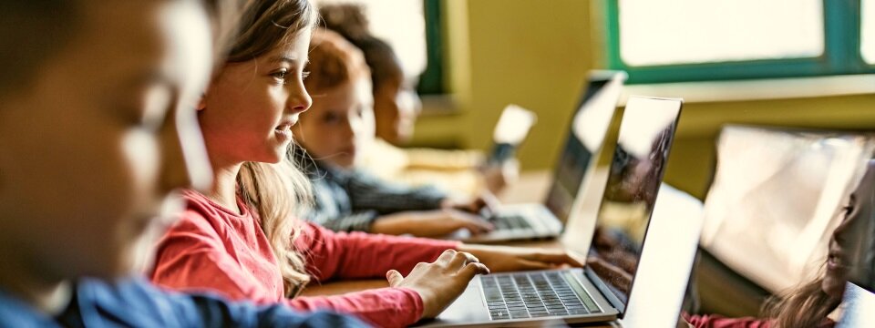 Vier Kinder sitzen an Tischen in einem Klassenzimmer und schauen auf Laptops.