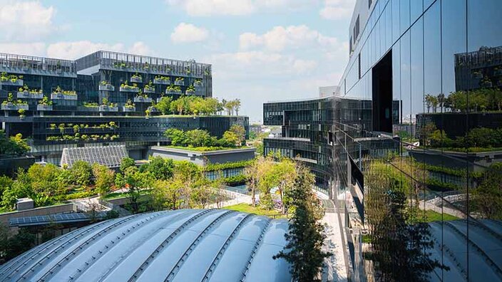 Moderne Gebäude mit viel grünem Bewuchs, einer Glasfront, in der sich der Himmel spiegelt