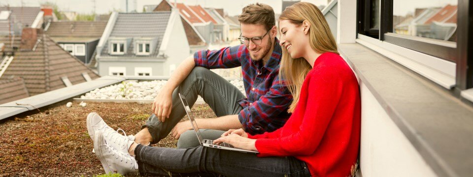 Eine junge Frau in einem roten Pullover sitzt mit einem jungen Mann auf einer Dachterrasse Sie sprechen miteinander und sie hält einen Laptop in der Hand.
