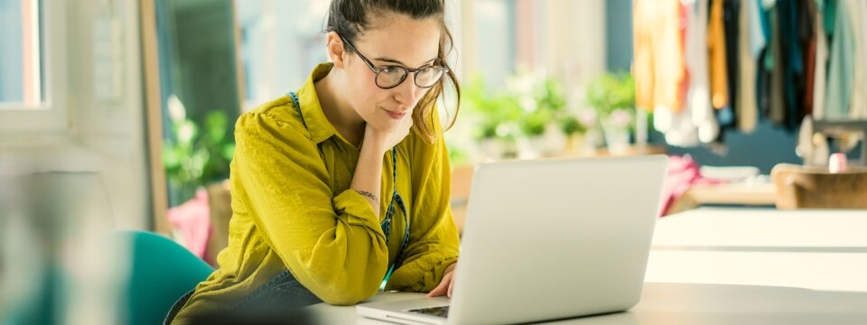 Eine Frau sitzt in ihrem Mode-Atelier an einem Schreibtisch und arbeitet an einem Laptop.