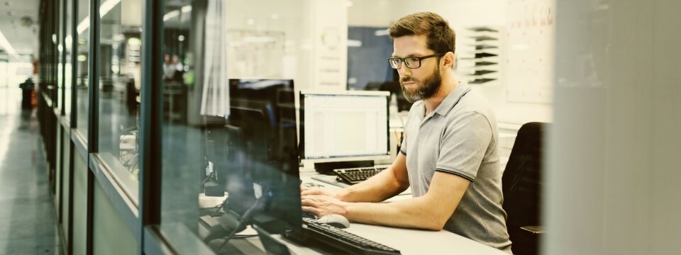 Ein Mann sitzt in einem Büro an einem Schreibtisch, schaut auf einen Monitor und arbeitet.