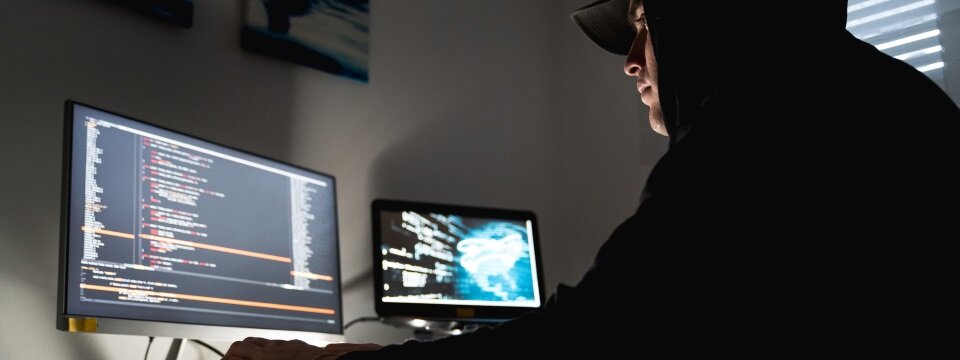 Ein Hacker mit einer schwarzen Kapuze sitzt an einem Schreibtisch und schaut sich Daten auf einem Monitor an.