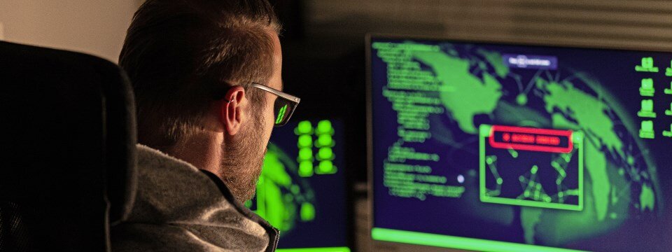 Ein Cyberkrimineller sitzt vor zwei Bildschirmen und plant seinen nächsten Phishing-Angriff auf ein Unternehmen.