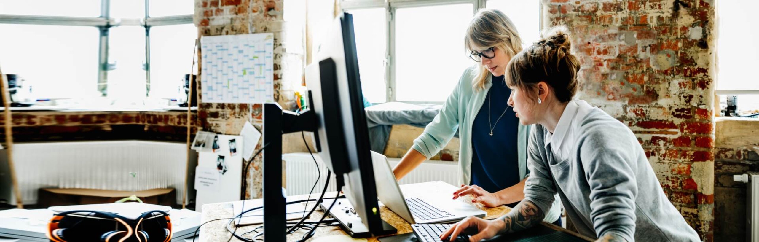 Zwei Frauen stehen an einem Hochtisch und arbeiten an einem Laptop und einem Computer.