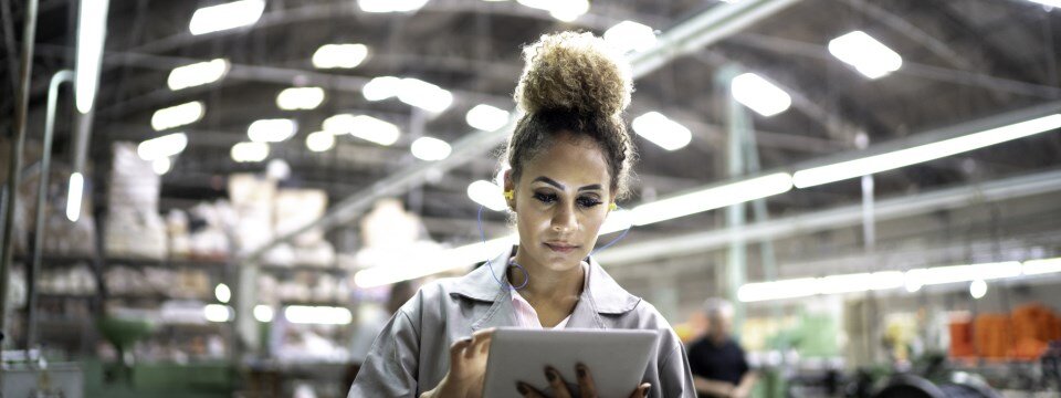 Frau mit Zopf guckt in einer gut beleuchteten Produktionshalle auf ihr Tablet in der Hand