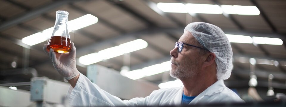 Mann hält in gut beleuchteter Halle chemische Probe im Glas in die Höhe, um sie zu prüfen