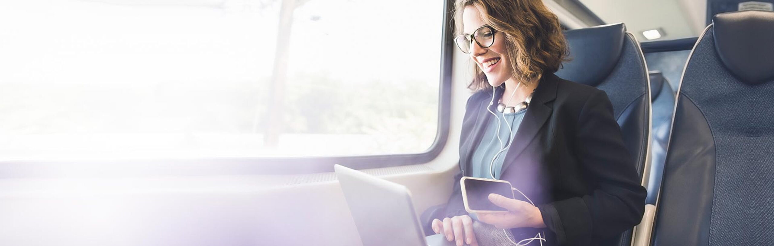 Eine Frau sitzt mit Notebook, Smartphone und Kopfhörern im Zug vor einem Fenster