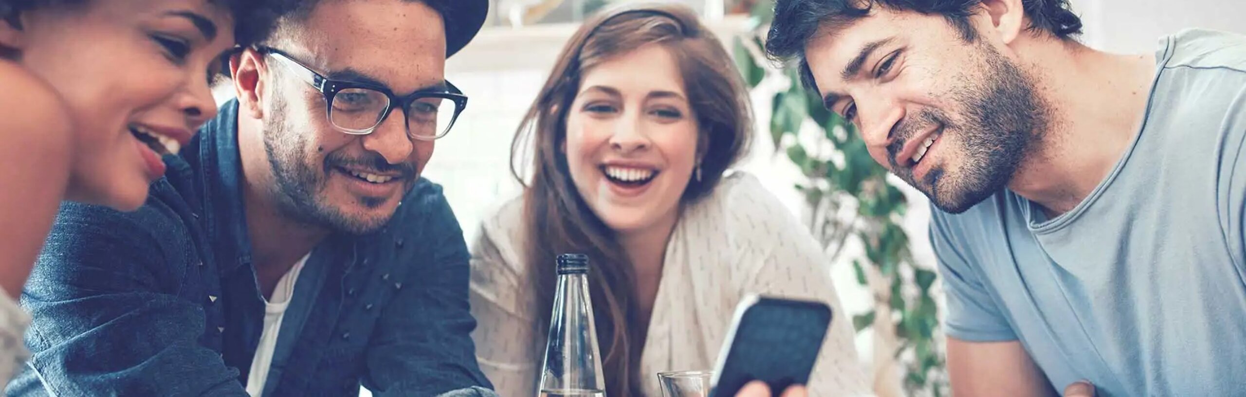 Vier junge Menschen sitzen um einen Tisch, gucken auf ein Smartphone und freuen sich.