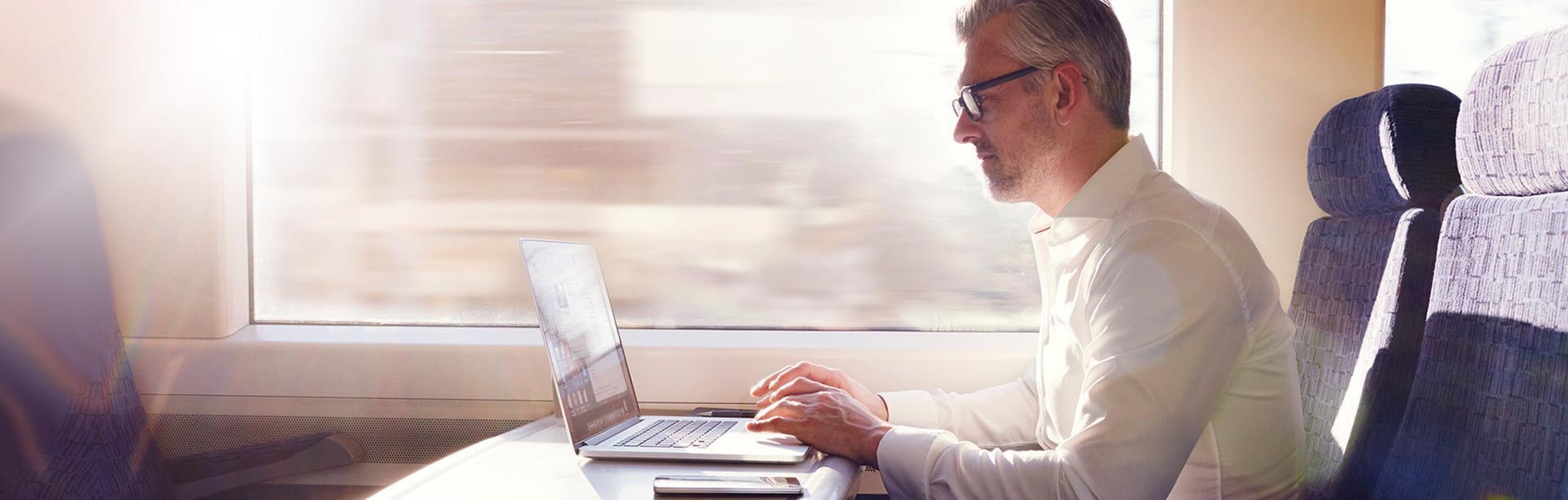 Ein Mann im weißen Hemd im Zug mit Laptop und Smartphone auf dem Tisch