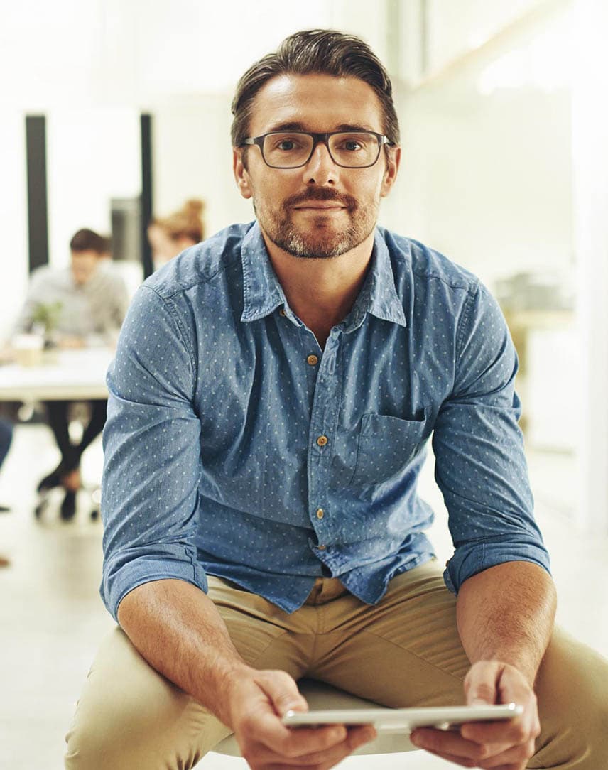 Ein Mann auf einem Bürodrehstuhl in einem Büro, der ein Tablet in der Hand hält und in die Kamera guckt.