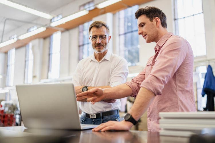 Zwei Männer stehen an einem Tisch mit Laptop und besprechen etwas.