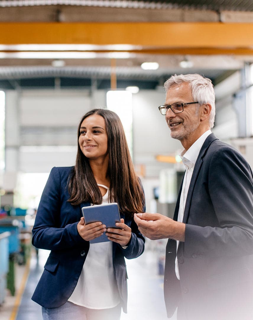 Eine Geschäftsfrau mit Tablet und ein Geschäftsmann besprechen etwas in einer Produktionshalle.