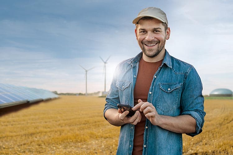 Ein Mann mit Kappe und Jeanshemd, steht auf einem Feld, hält ein Smartphone in der Hand und lächelt, im Hintergrund eine PV-Anlage, Windräder und eine Biogasanlage