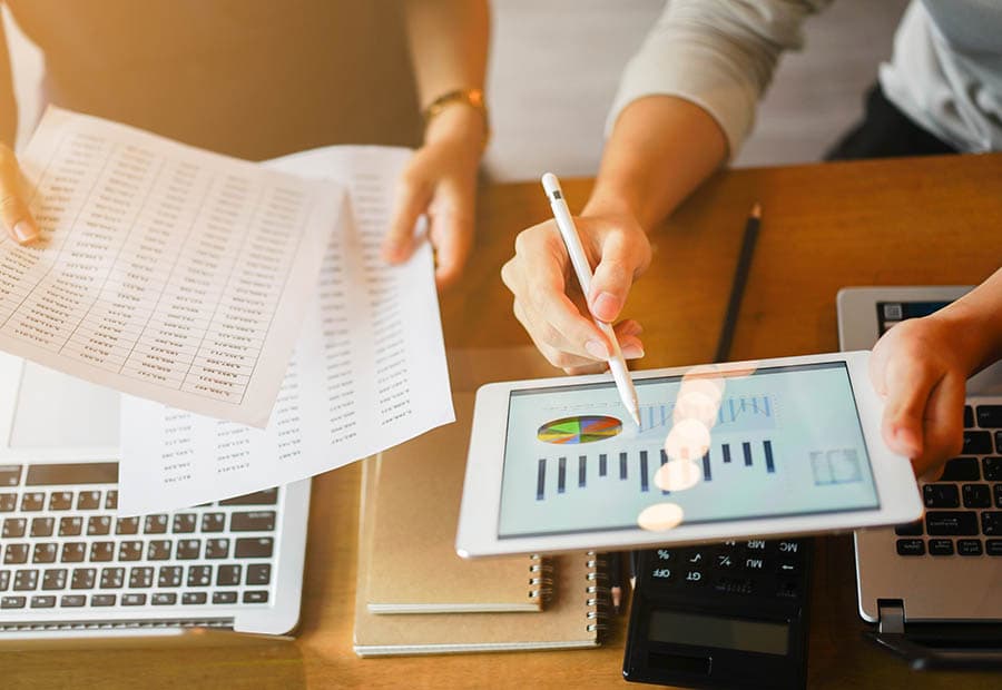 Ein Mann zeigt auf ein Diagramm auf einem Tablet, daneben hält eine Frau Zettel mit Damen über einem Laptop