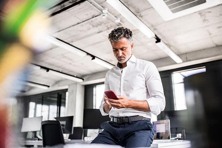 Ein Mann im weißen Hemd sitzt an einen Schreibtisch gelehnt und guckt etwas auf seinem Handy an.