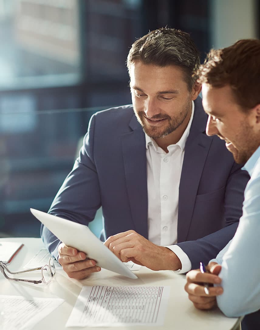 Ein Mann im Anzug und ein weiterer Mann in einem hellblauen Hemd sitzen an einem Tisch und schauen auf ein Tablet, vor ihnen liegen Unterlagen und Stifte.