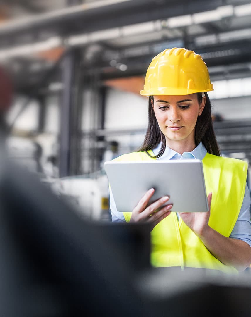 Eine Frau mit Warnweste und Helm arbeitet mit dem Tablet in einer Industriehalle.