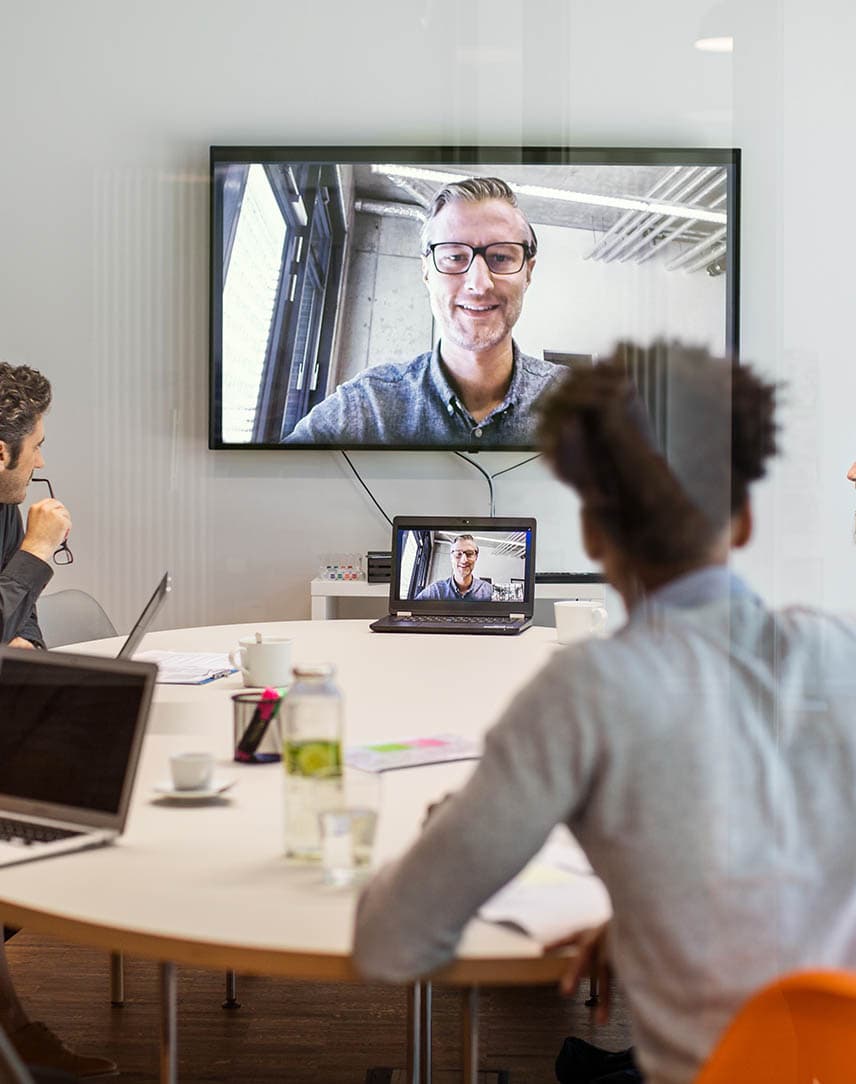 In einem Besprechungsraum sitzen Frauen und Männer und nehmen an einer Videokonferenz mit einem Kollegen teil.