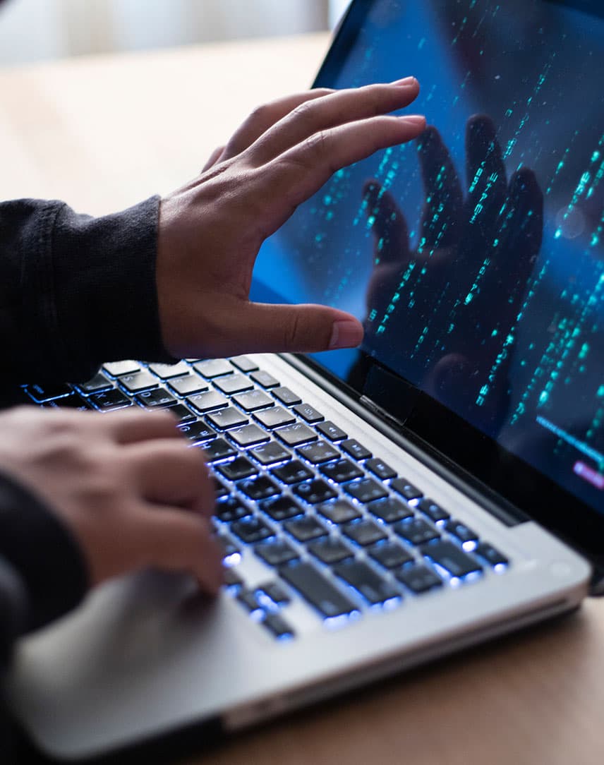 Ein Hacker sitzt an einem Laptop und zeigt mit seiner Hand auf den Bildschirm. Auf dem Bildschirm sind Zahlen und Buchstaben zu sehen.