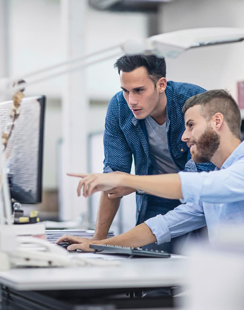 Zwei junge Männer am Schreibtisch in einem Büro. Einer der Männer sitzt auf seinem Stuhl und zeigt mit seinem Arm auf seinen Monitor. Der andere Mann steht neben ihm und schaut auf den Monitor.