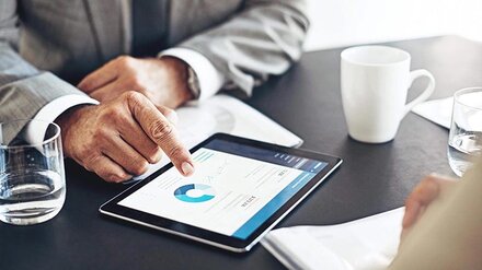 Licht-Finanzierung Zwei Personen betrachten ein Kreisdiagramm auf einem Tablet, das auf einem Besprechungstisch liegt.