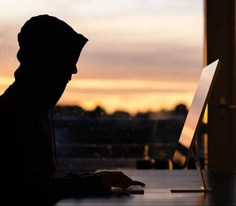 DDoS-Schutz-Angriff Stilisierter Hacker mit Kapuze über dem Kopf sitzt vor einem Computer