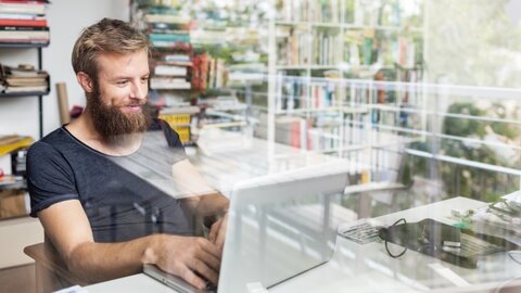 Optimale Bandbreite für das Home Office Ein bärtiger Mann sitzt an seinem Home-Office-Arbeitsplatz und lächelt.