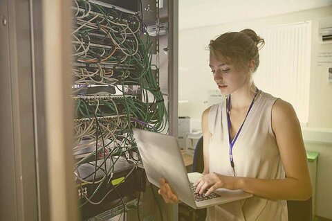 Passende IT für die Schule Eine Frau arbeitet mit Laptop hinter einemServerschrank.