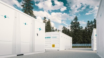 Batteriespeicher Im Vordergrund weiße Batteriespeicher, im Hintergrund Bäume, ein Windrad und eine PV-Anlage